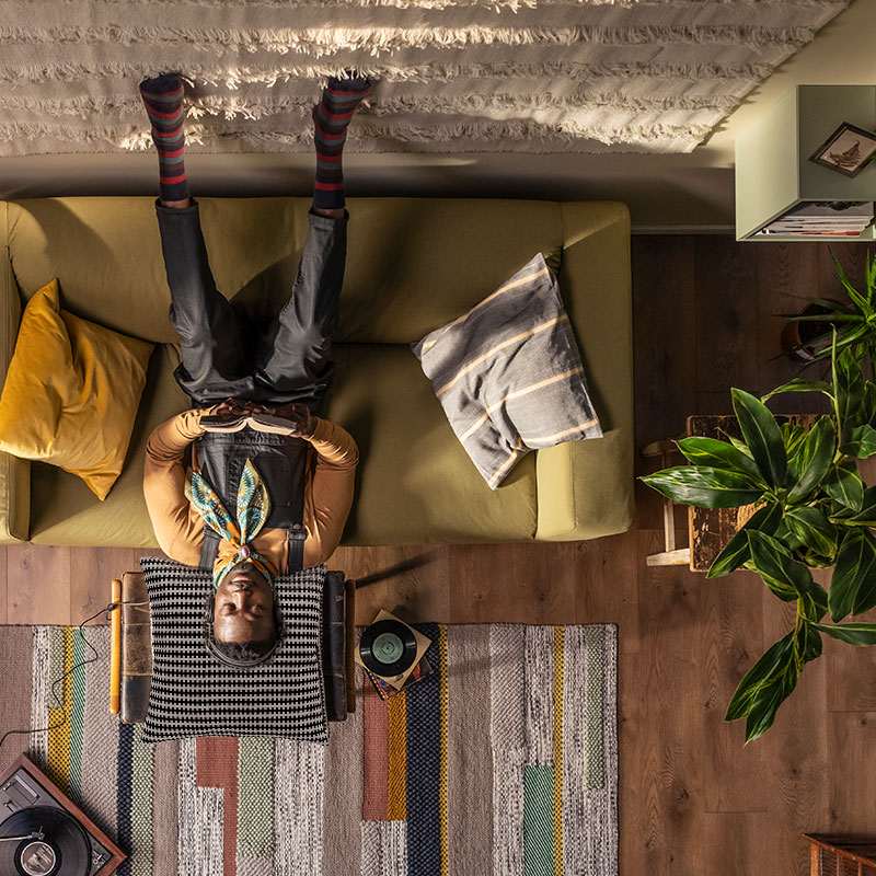 Vista aérea: persona con orejas de conejo, boca abajo en sofá verde, sala acogedora con plantas, guitarra y tocadiscos.