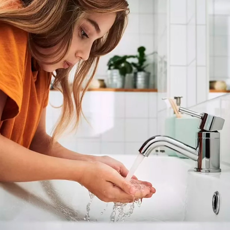 Niña se lava las manos en un lavabo moderno con agua corriente, en un baño luminoso con azulejos y plantas al fondo.