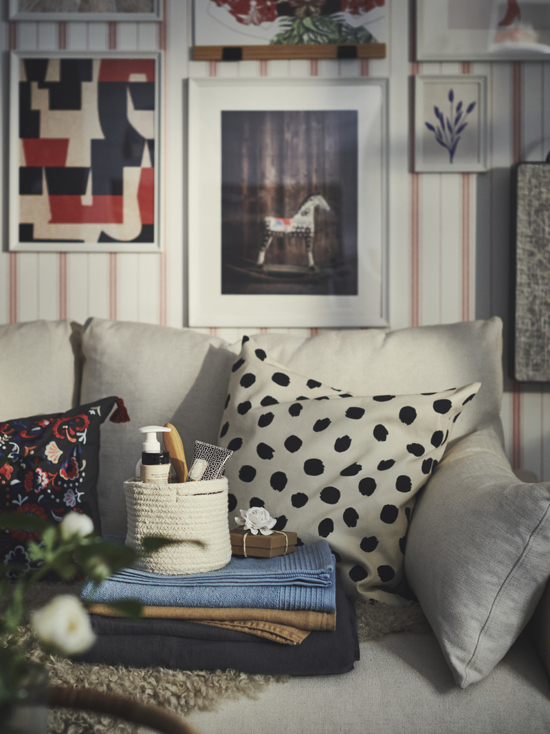 Sofa with patterned cushions, folded blankets, and basket with items, in front of wall decorated with pictures. Cozy atmosphere.