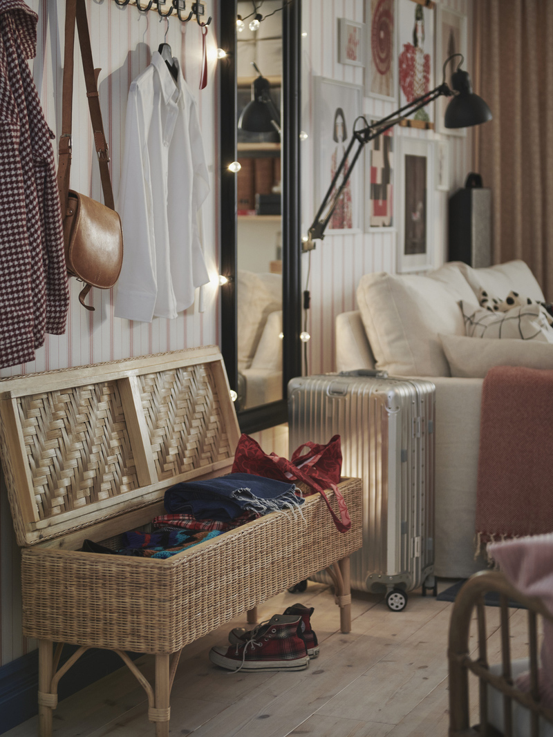 Cozy entryway with a wicker storage bench, coat rack with clothes, and illuminated full-length mirror.