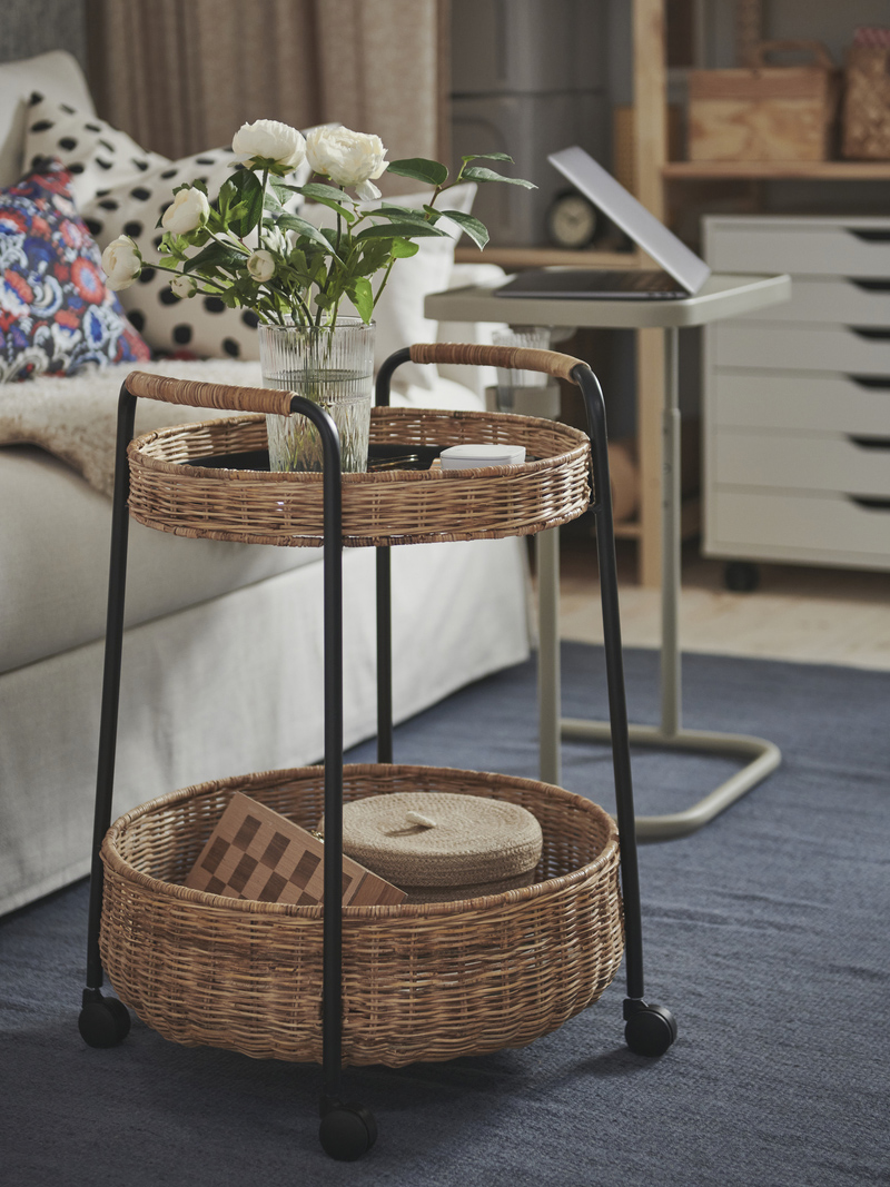 Round wicker side table with wheels and white flowers, on a blue rug, next to a sofa and study area.