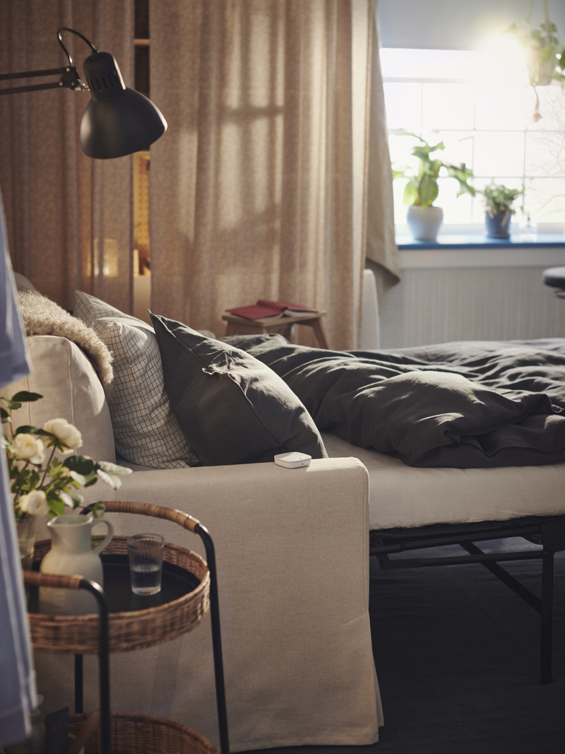 Bed with gray bedding, nightstand with flowers, warm window light, and sheer curtains. Serene atmosphere.