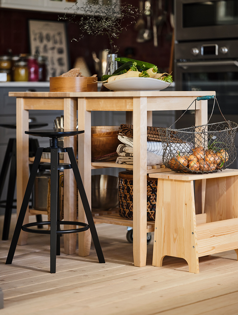 Tall pine tables with food to prepare on top, with black high stool and a bench underneath there is a wire basket with food to prepare.