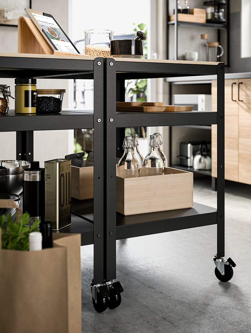 Carro de cocina alto, debajo tiene cajas de madera con botellas dentro, encima hay una tabla gráfica con una tabla de cortar y tarros con tapas.