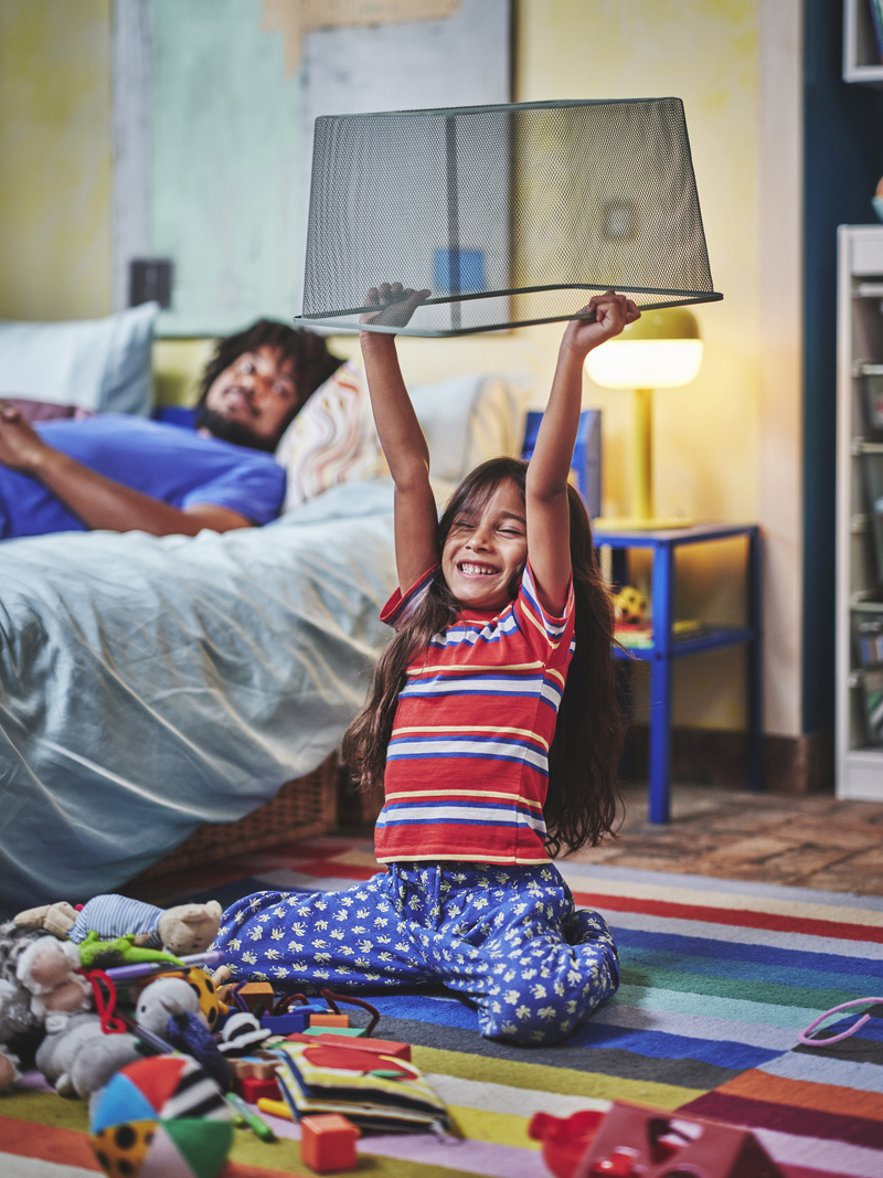 Smiling girl plays with a lampshade, while an adult rests on the bed. A playful and relaxed ambiance in a bedroom.