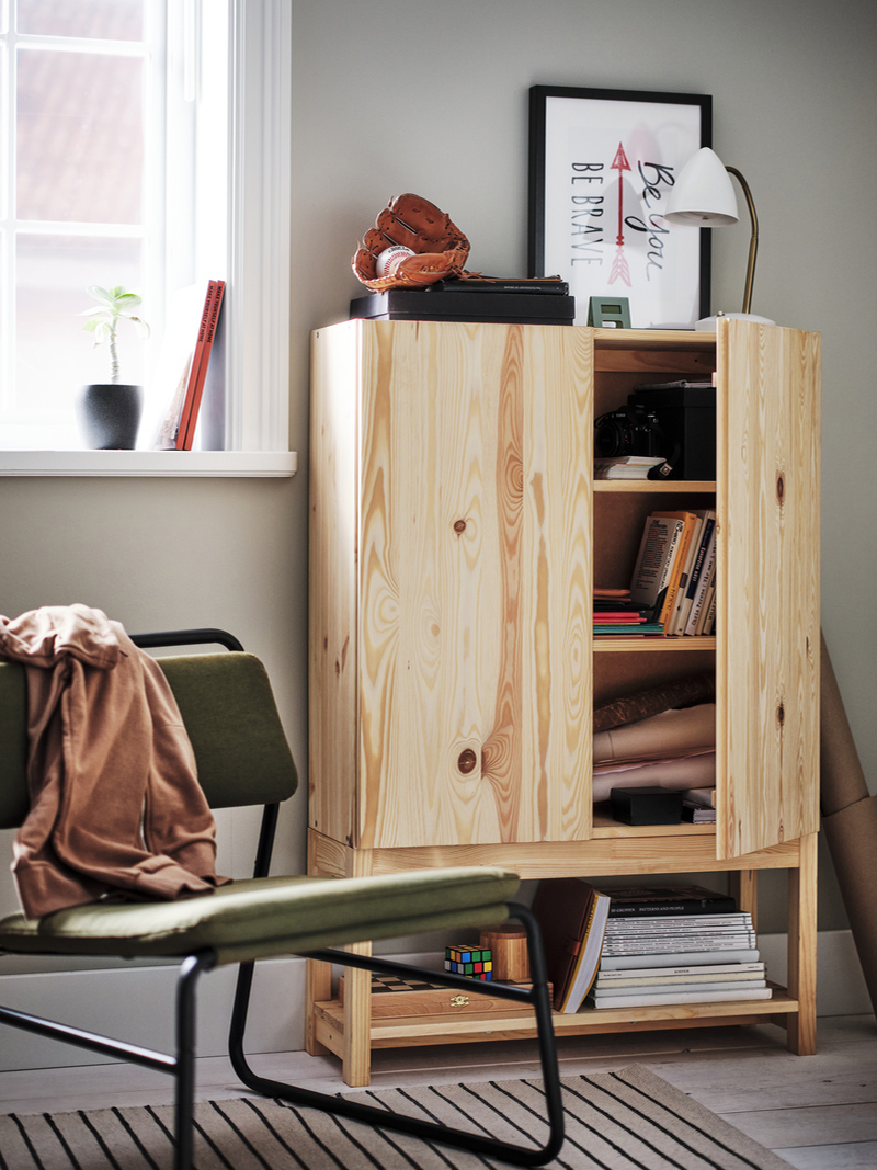 Light wood cabinet with books and objects, next to a green chair and a white lamp. A tidy and cozy ambiance in a bright room.