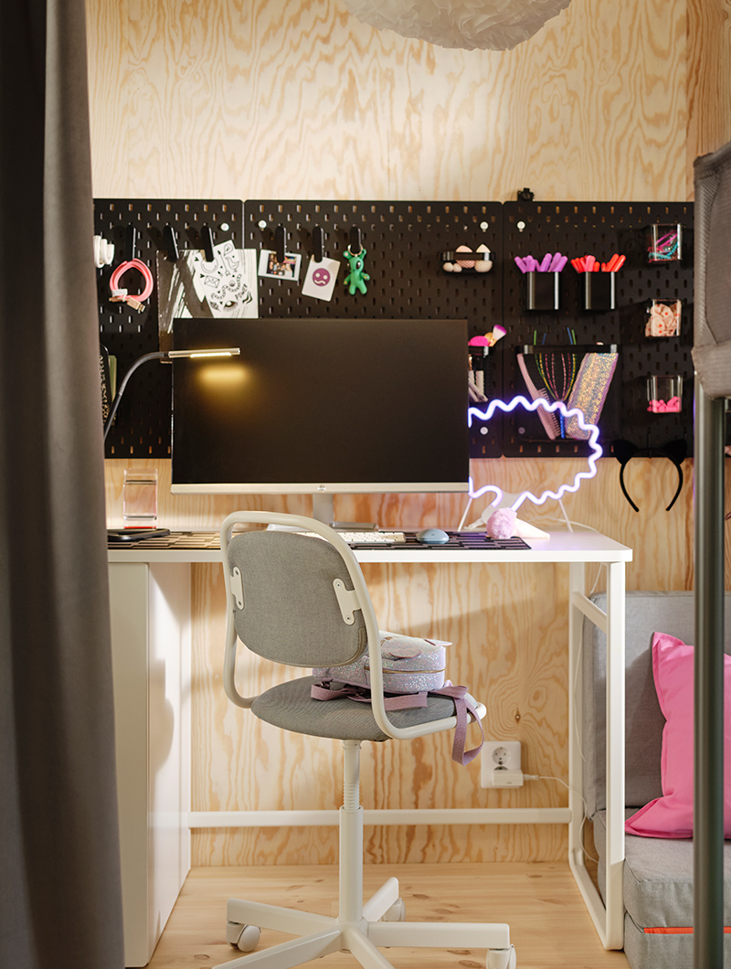 Un escritorio blanco con monitor, silla y un tablero perforado negro lleno de accesorios. Un ambiente de estudio moderno y funcional en un cuarto juvenil.