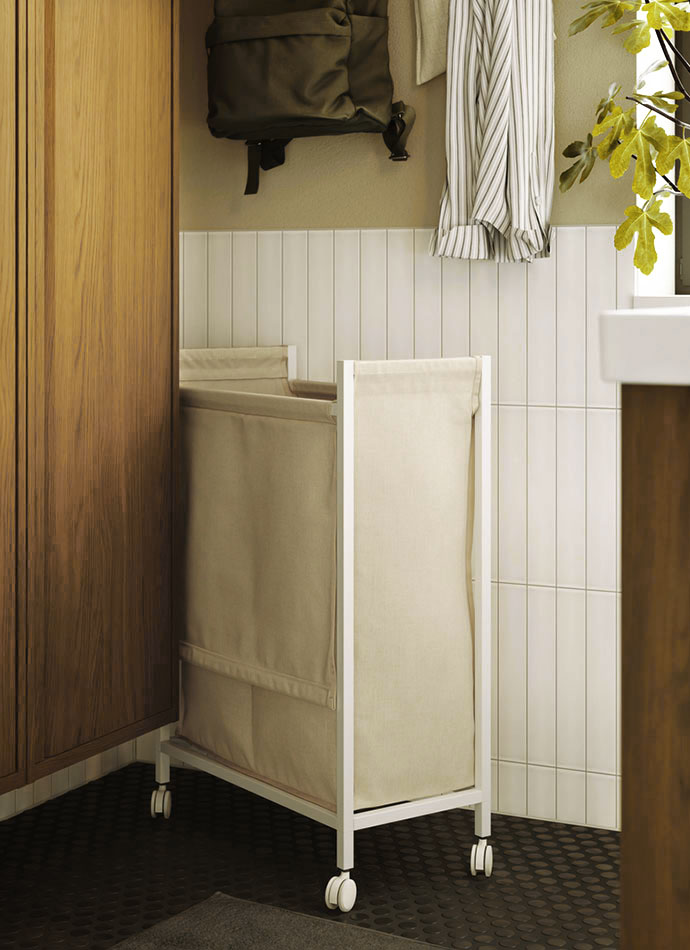 Cesto de ropa beige con ruedas en esquina de lavandería con paneles de madera, azulejos blancos y detalles verdes.