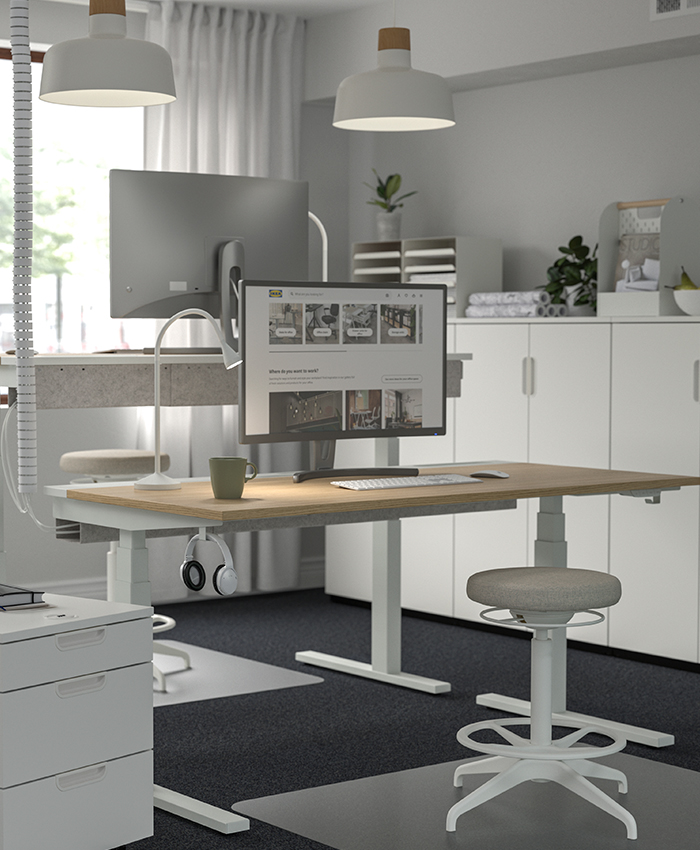Oficina moderna con escritorios regulables, monitores, taburete ergonómico y almacenaje blanco bajo iluminación suave.