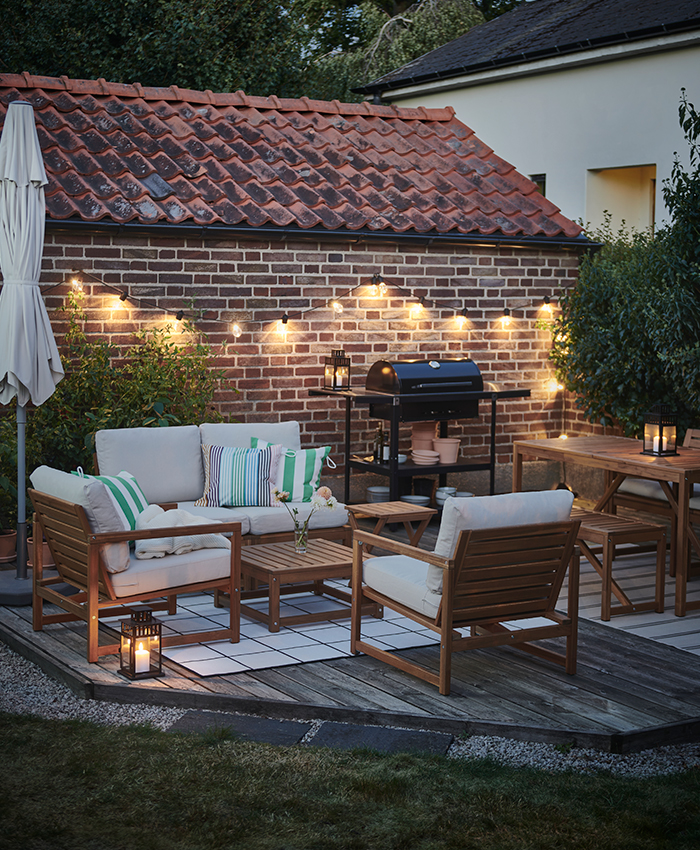 Zona exterior acogedora con muebles de jardín de madera y cojines blancos, guirnalda de luces cálidas, barbacoa negra y mesa de comedor, todo sobre una tarima de madera junto a una pared de ladrillo.