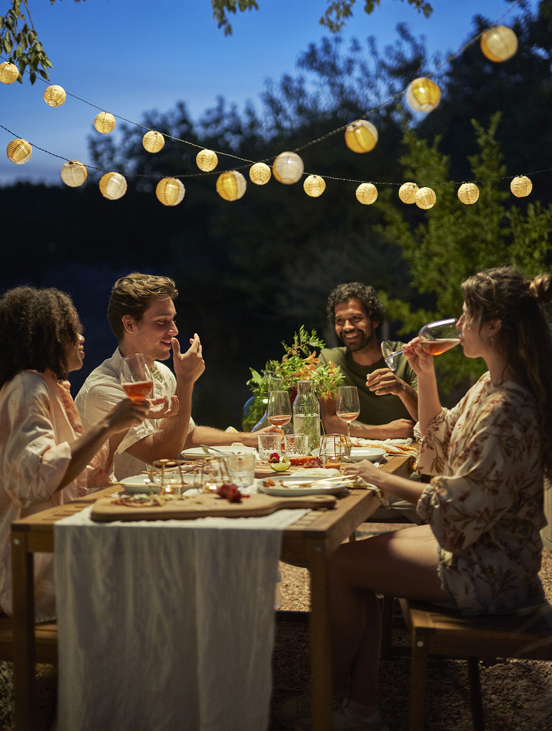 Four friends enjoy an outdoor dinner under warm string lights. The ambiance is jovial, with wine and food on the wooden table.