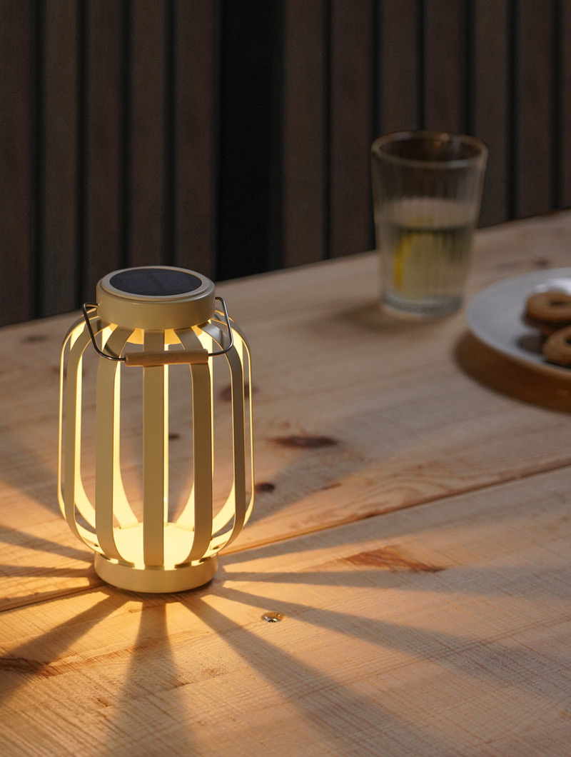 A white solar lantern illuminates a wooden table. A glass of water and cookies create a tranquil and cozy evening ambiance.