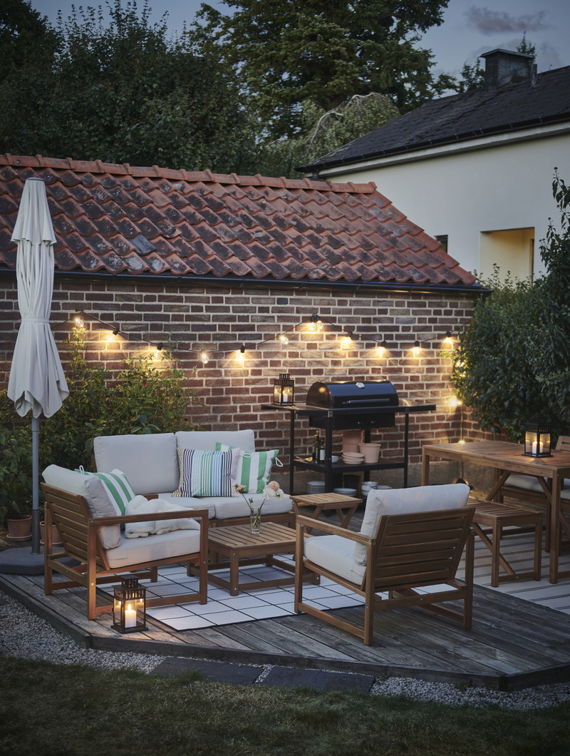 Patio lit with warm string lights, light wood furniture, and green and white cushions. A cozy ambiance for outdoor evenings.