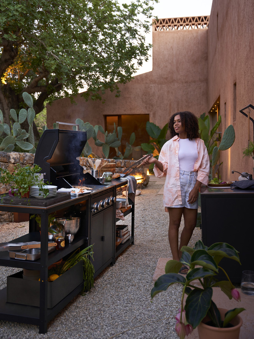 Woman cooking on an outdoor grill in a patio with plants and earthy-toned walls. A warm and relaxed ambiance.