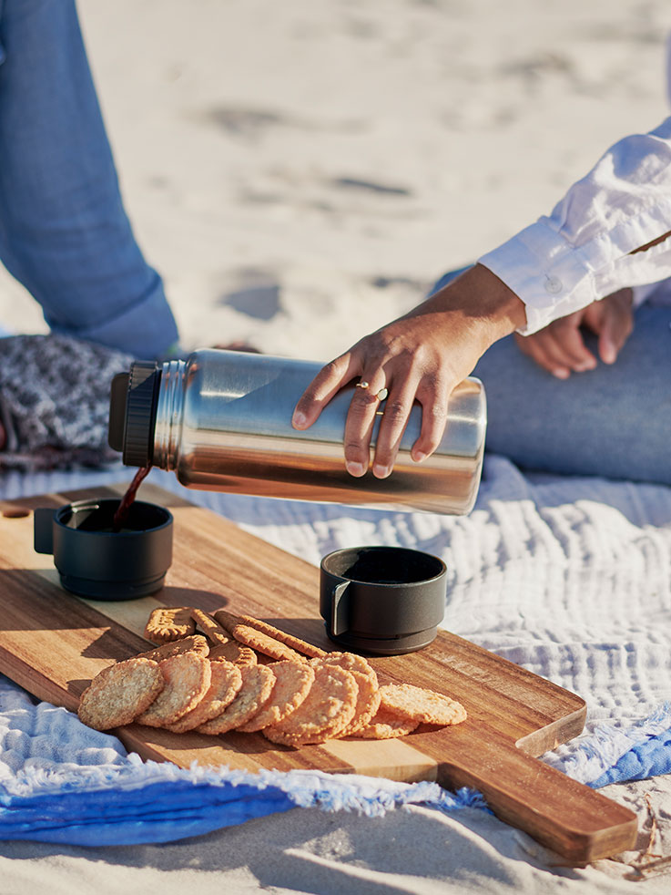 Persona sirviendo café de un termo en una playa, con galletas sobre tabla de madera. 