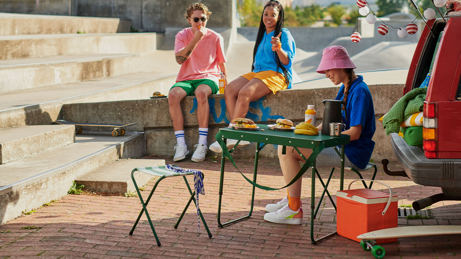 Young people sharing food at a skate park. Green table, orange cooler, relaxed and sunny atmosphere.