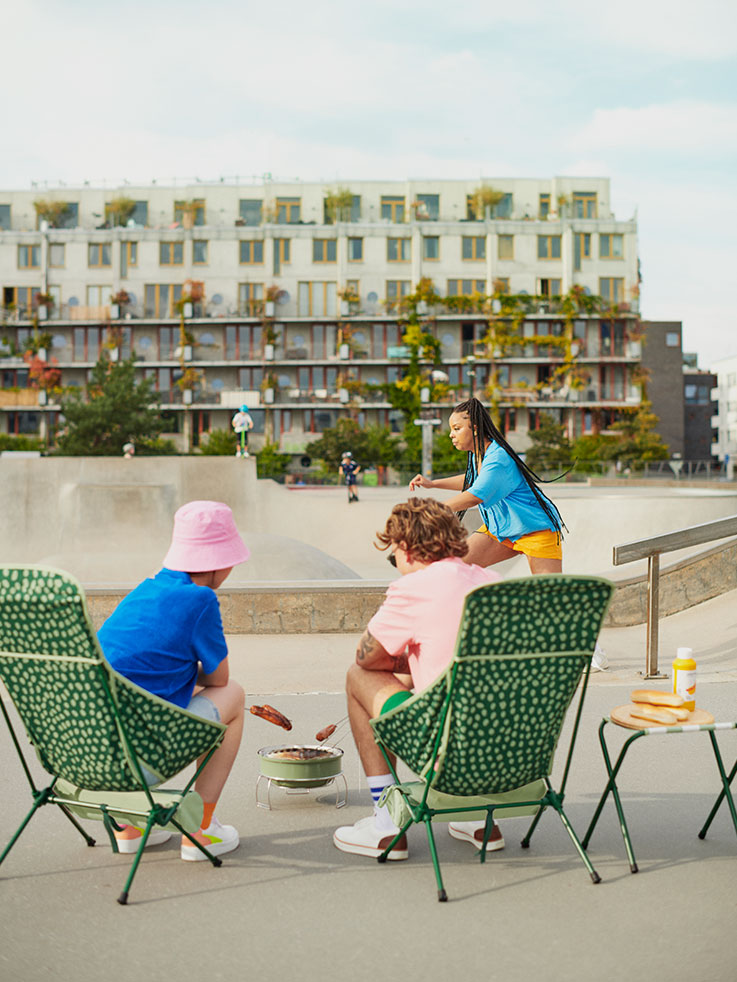 Friends relaxing in green folding chairs, with barbecue at urban skatepark. Casual and modern atmosphere.