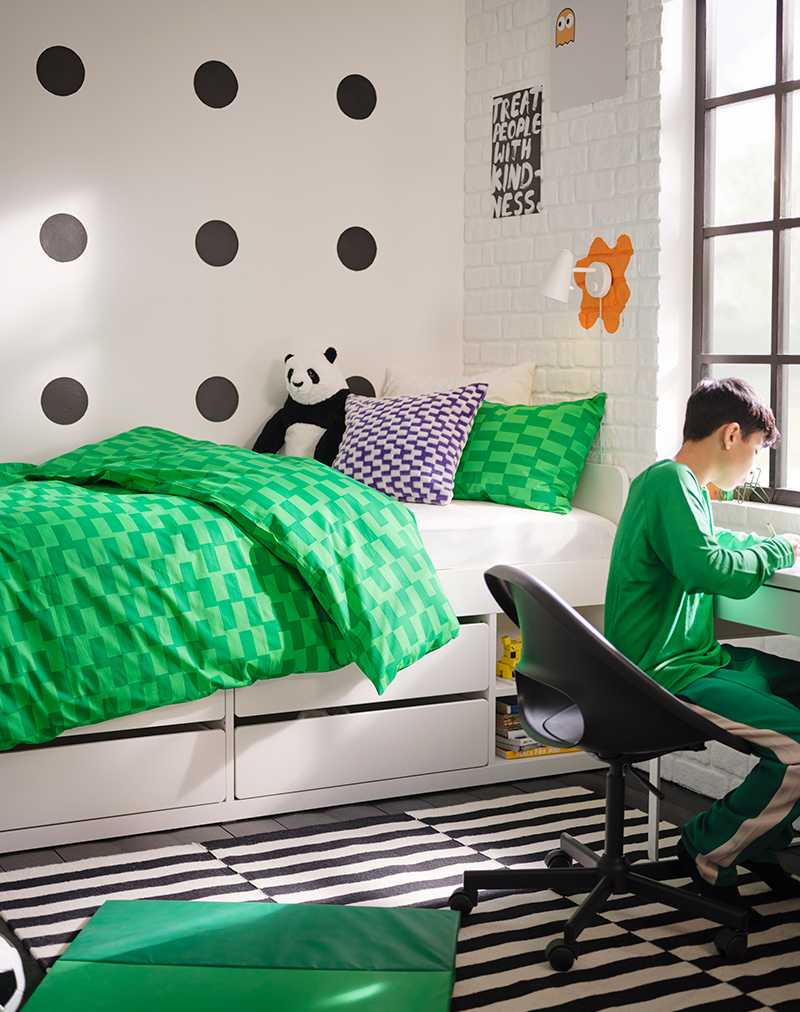Una cama SLAKT cubierta con un juego de edredón verde BLÅSKATA junto a un niño escribiendo