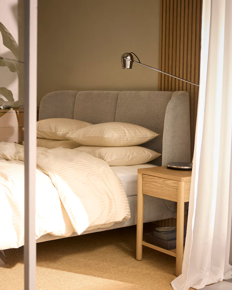 Bed with gray headboard, beige sheets, wooden nightstand, and metal lamp. Serene, minimalist atmosphere.