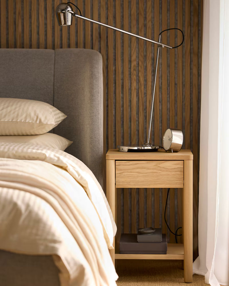 Light wood nightstand next to gray bed, with modern lamp and alarm clock. Warm and functional atmosphere.