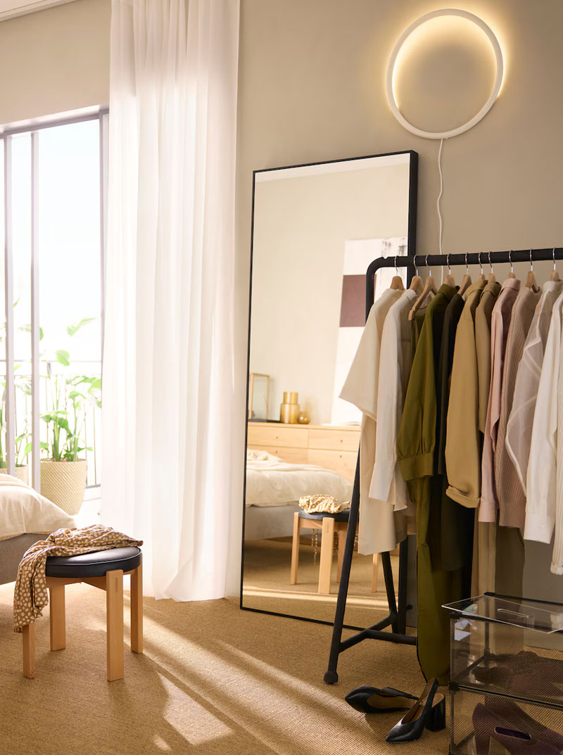 Bedroom with clothes rack, full-length mirror, wooden stool, and window. Bright atmosphere.
