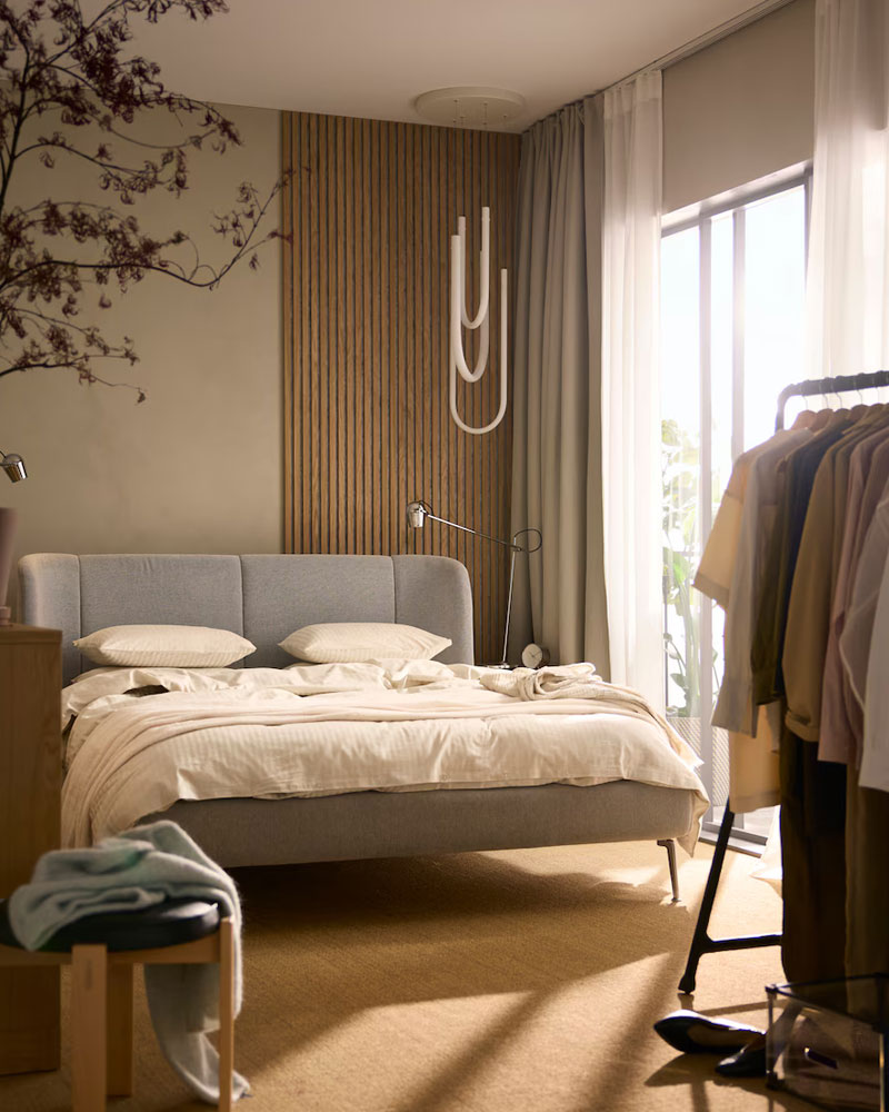 Bedroom with gray bed, wooden wall, decorative branches, and clothes rack. Bright and serene atmosphere.