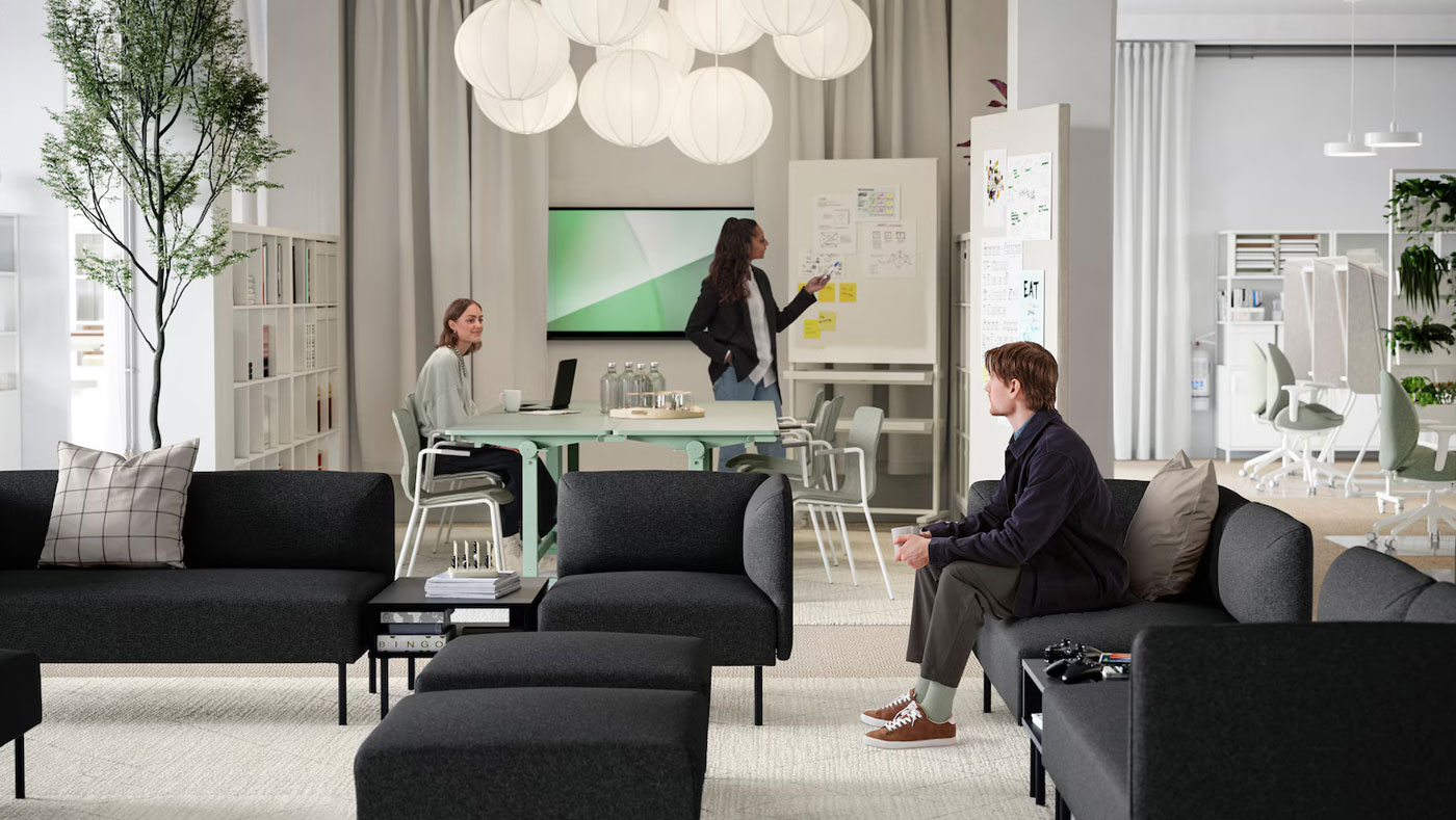Amplia sala de espera de una oficina moderna y creativa, al fondo se ve una mesa de reuniones con alguien exponiendo, el lugar parece espacioso, luminoso y elegante.