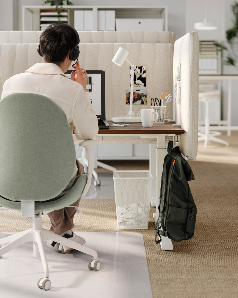 Plano posterior de una persona sentada en una silla de trabajo en un cubículo de oficina, incluso se puede ver un protector de suelo para las ruedas de la silla.