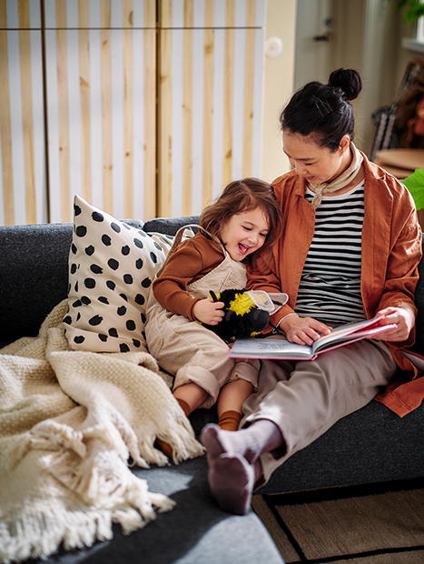 Mother and daughter reading together on the sofa, with a blanket and decorative cushion in a cozy setting.
