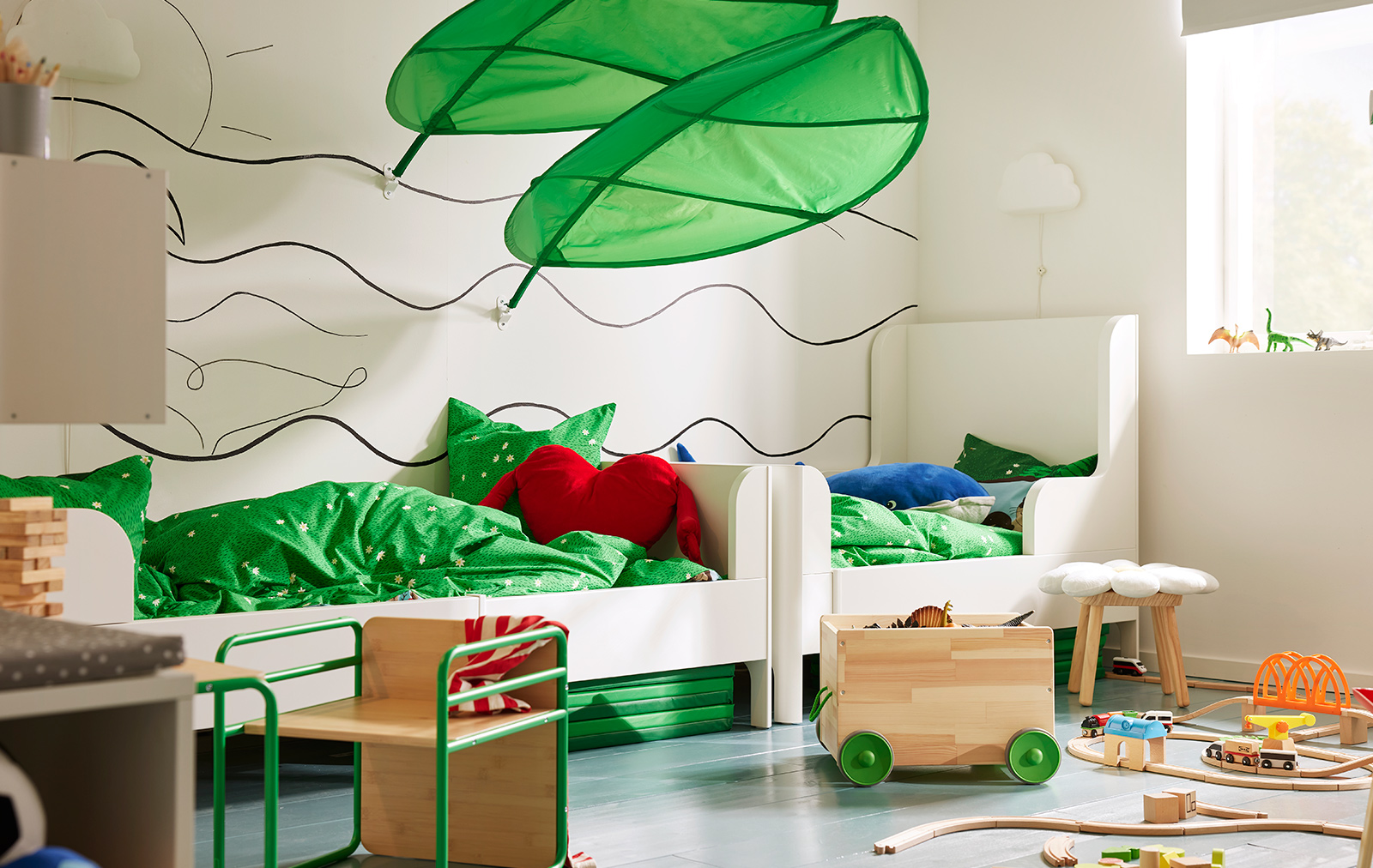 Habitación infantil con dos camas blancas, ropa de cama verde, cojín rojo y decoraciones de hojas.
