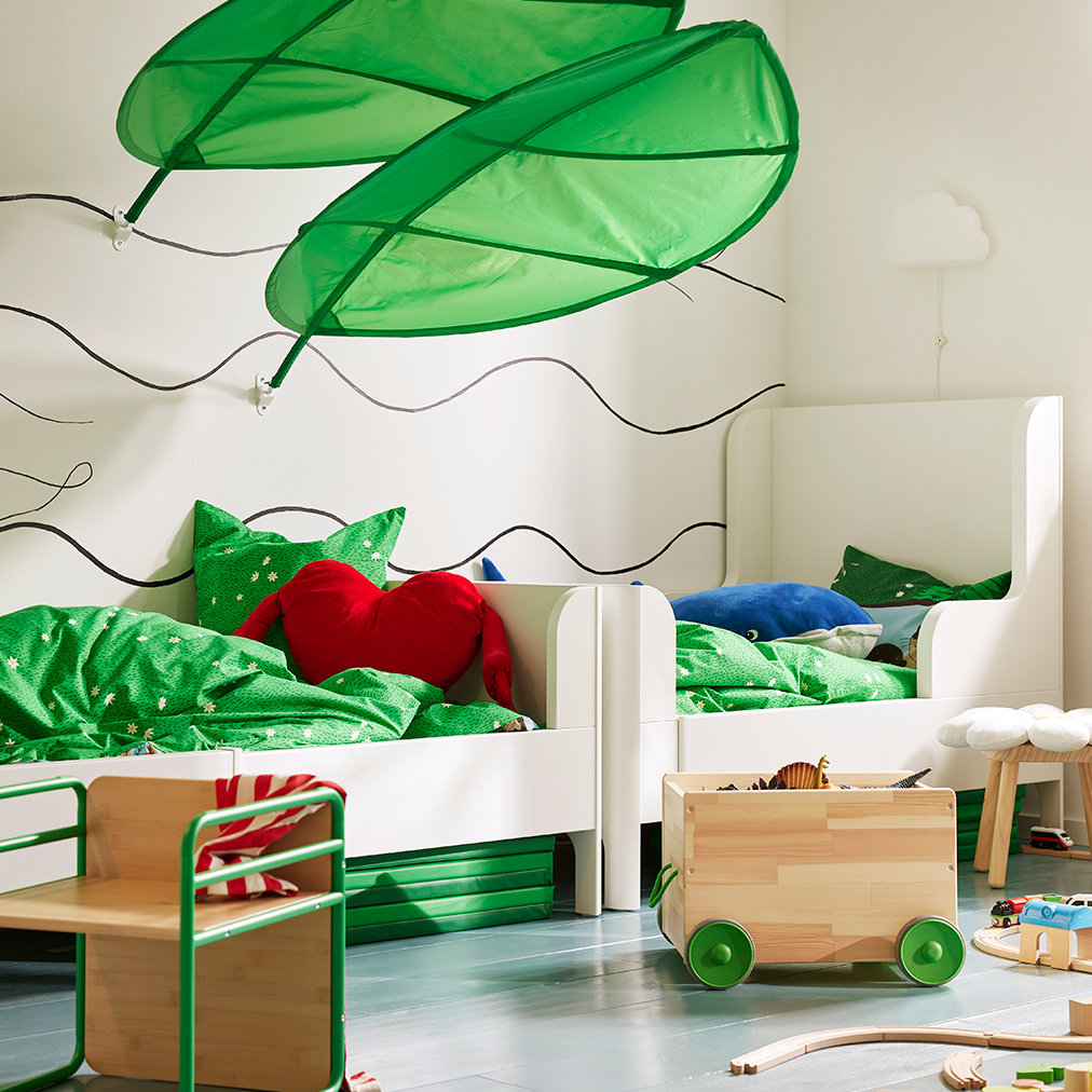 Habitación infantil con dos camas blancas, ropa de cama verde, cojín rojo y decoraciones de hojas.