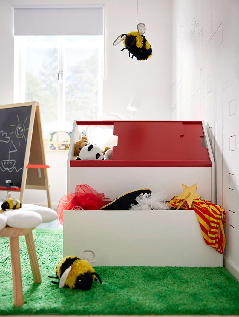Caja de juguetes blanca y roja, con peluches, en cuarto infantil. Abeja de peluche colgando. Ambiente lúdico.