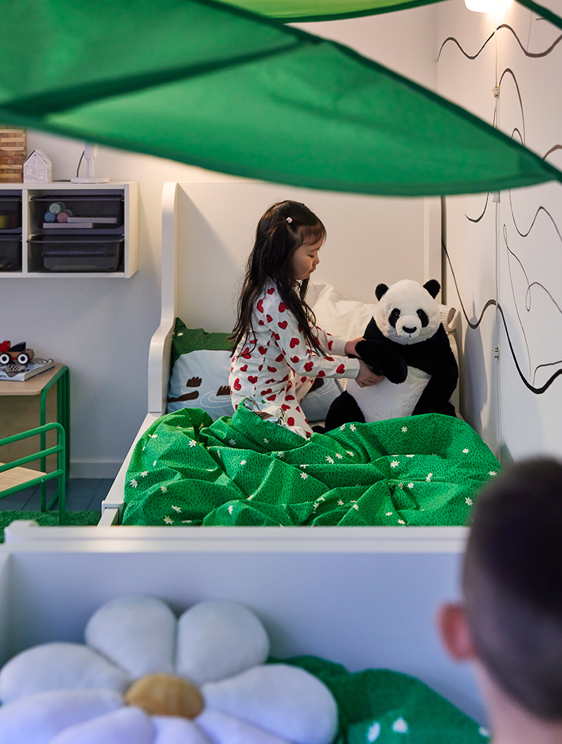 Niña en pijama con panda de peluche en cama blanca y verde bajo toldo hoja. Ambiente infantil.