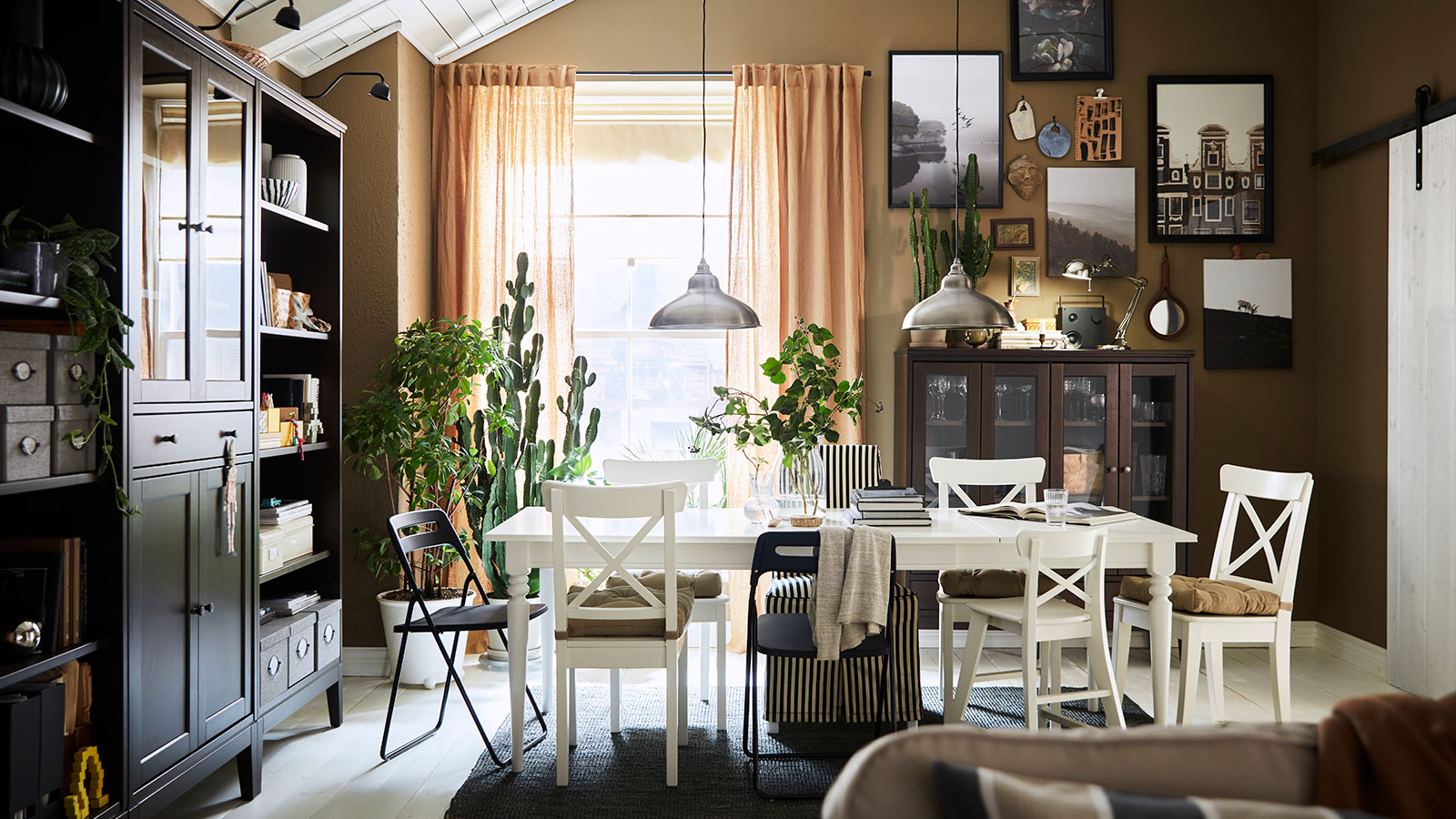 Comedor de estilo tradicional con toques vintage, la mesa es blanca, las sillas con cojines, hay un gran ventanal con cortinas beige por donde entra la luz, y en la pared de la derecha hay una estantería y vitrina, al fondo una vitrina.