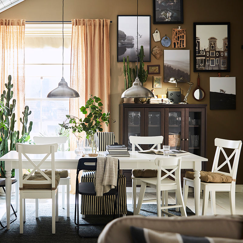 Comedor de estilo tradicional con toques vintage, la mesa es blanca, las sillas con cojines, hay un gran ventanal con cortinas beige por donde entra la luz, y en la pared de la derecha hay una estantería y vitrina, al fondo una vitrina.