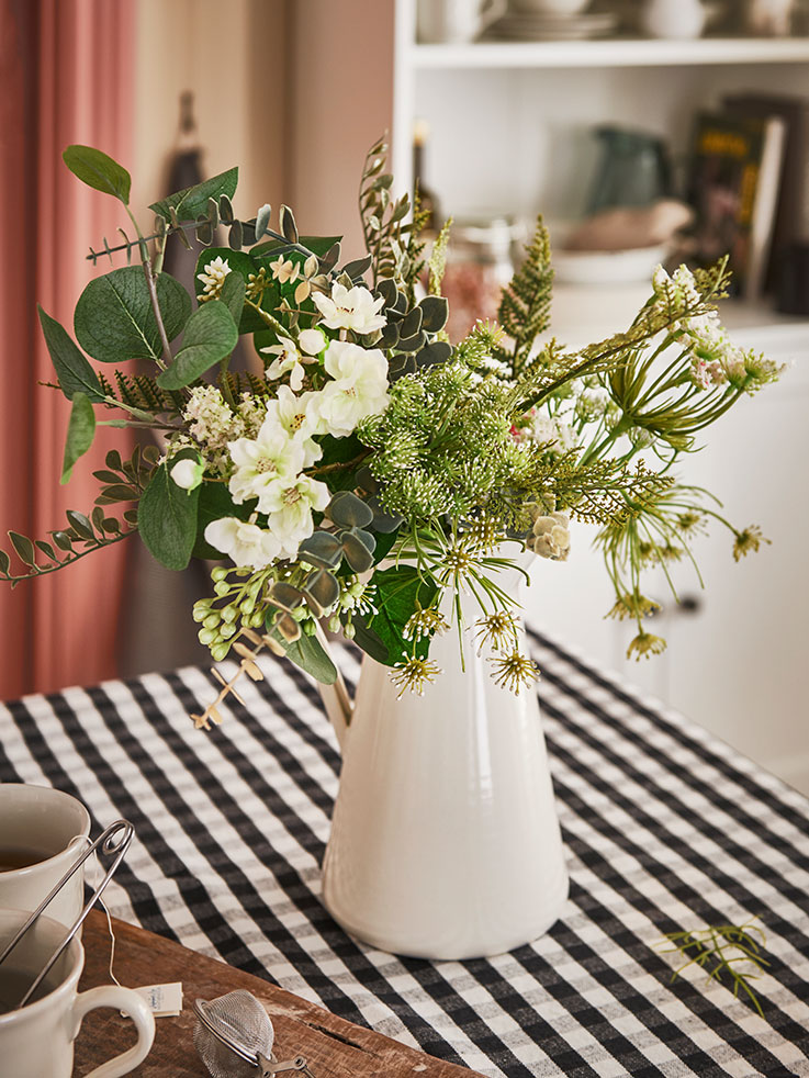 Florero blanco en forma de jarrón sobre una mesa de estilo tradicional con un mantel a cuadros blancos y negros.