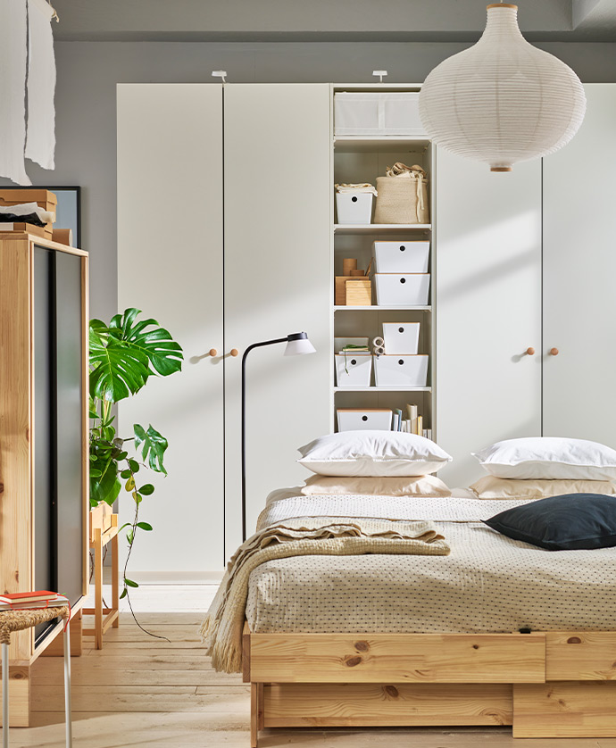 Bedroom with light wood bed, white PAX wardrobes with open storage and boxes, paper pendant lamp, and decorative plant.