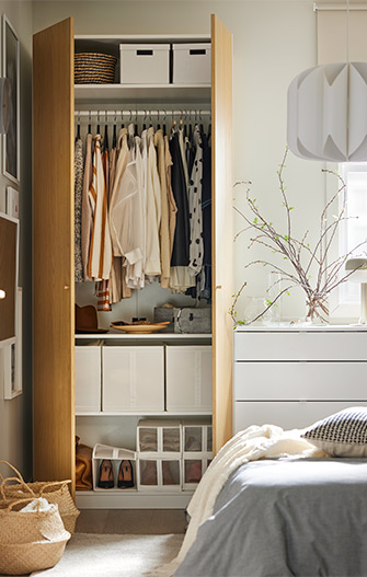 Open wardrobe with wooden doors, neatly hung clothes, organized boxes and shoes; next to a white chest of drawers and natural decor.
