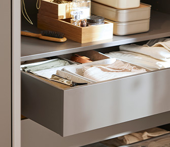 Open drawer with inner organizers and folded clothes, with trays and boxes on the shelf above.