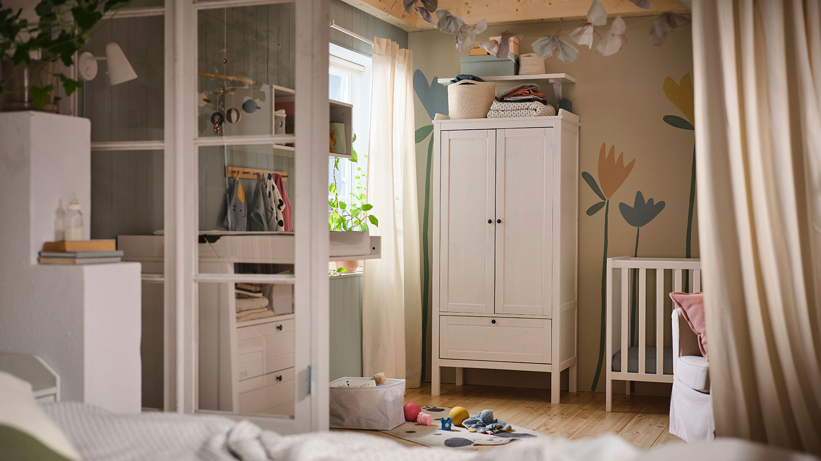 Una habitación de bebé, un cambiador frente a una ventana, una estantería modular, un armario junto a una cuna y juguetes en el suelo junto a una cortina.