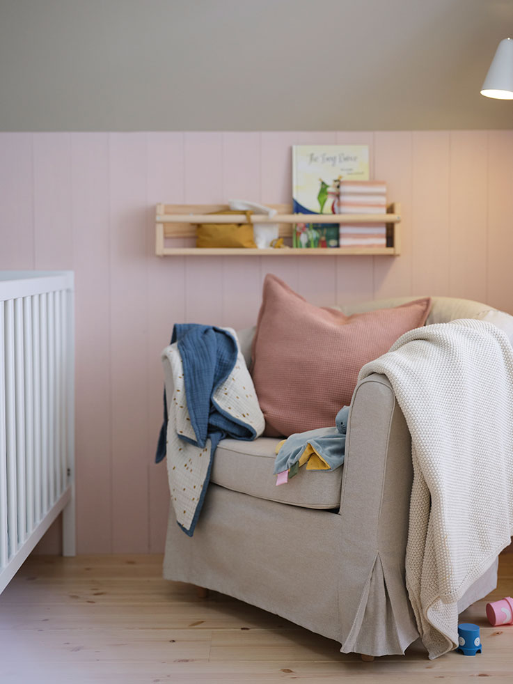 Sillón con funda textil con un cojín rosa y una colcha y una manta de bebé, hay una estantería de pino colgada en la pared y se ve parte de una cuna.