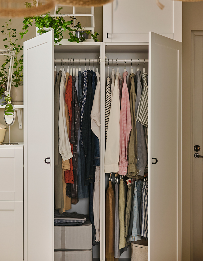 Armario blanco abierto con ropa colgada organizada por colores, cajas de almacenaje en la parte inferior y plantas decorativas en la parte superior.