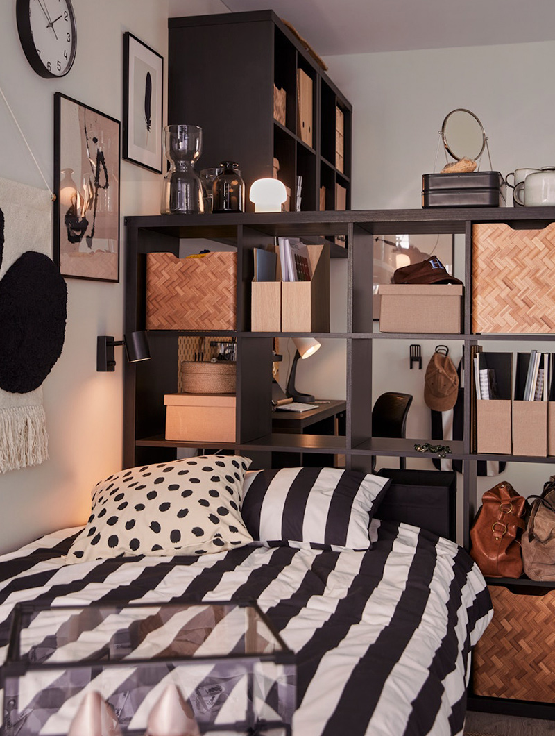 Dormitorio con cama de rayas blancas y negras, estantería oscura con adornos y escritorio.