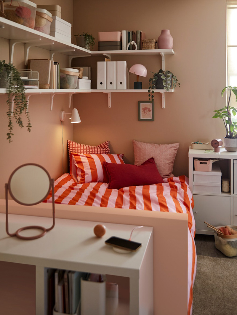 Dormitorio en tonos rosa y rojo, cama con rayas, estantes blancos y espejo. Ambiente juvenil.