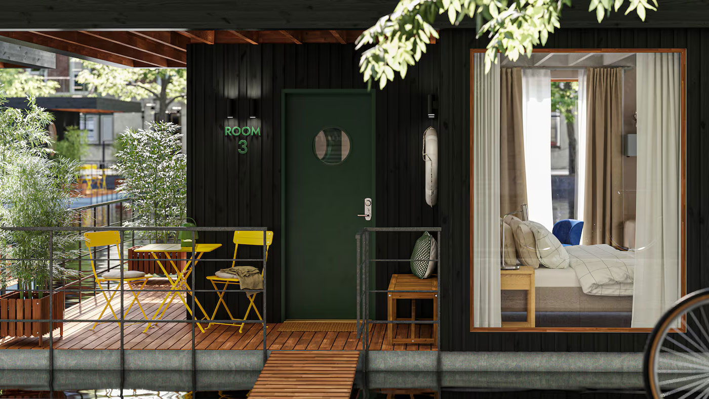 Vista frontal de una habitación flotante tipo hostal sobre un lago, con una amplia ventana que deja ver el dormitorio interior. Afuera, hay mesas y sillas amarillas de exterior que completan un ambiente acogedor sobre una plataforma flotante.