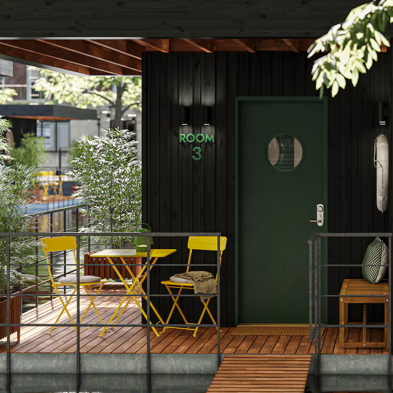 Vista frontal de una habitación flotante tipo hostal sobre un lago, con una amplia ventana que deja ver el dormitorio interior. Afuera, hay mesas y sillas amarillas de exterior que completan un ambiente acogedor sobre una plataforma flotante.