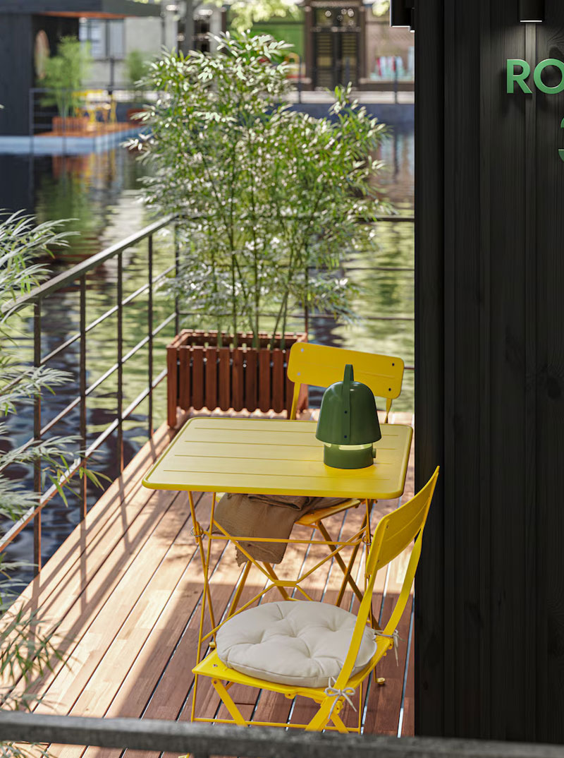 el exterior de la habitación del hostal, con un conjunto de sillas y mesa amarillas, hay un balcón metálico y una jardinera con finos bambúes, y al fondo las demás habitaciones y el agua del lago.