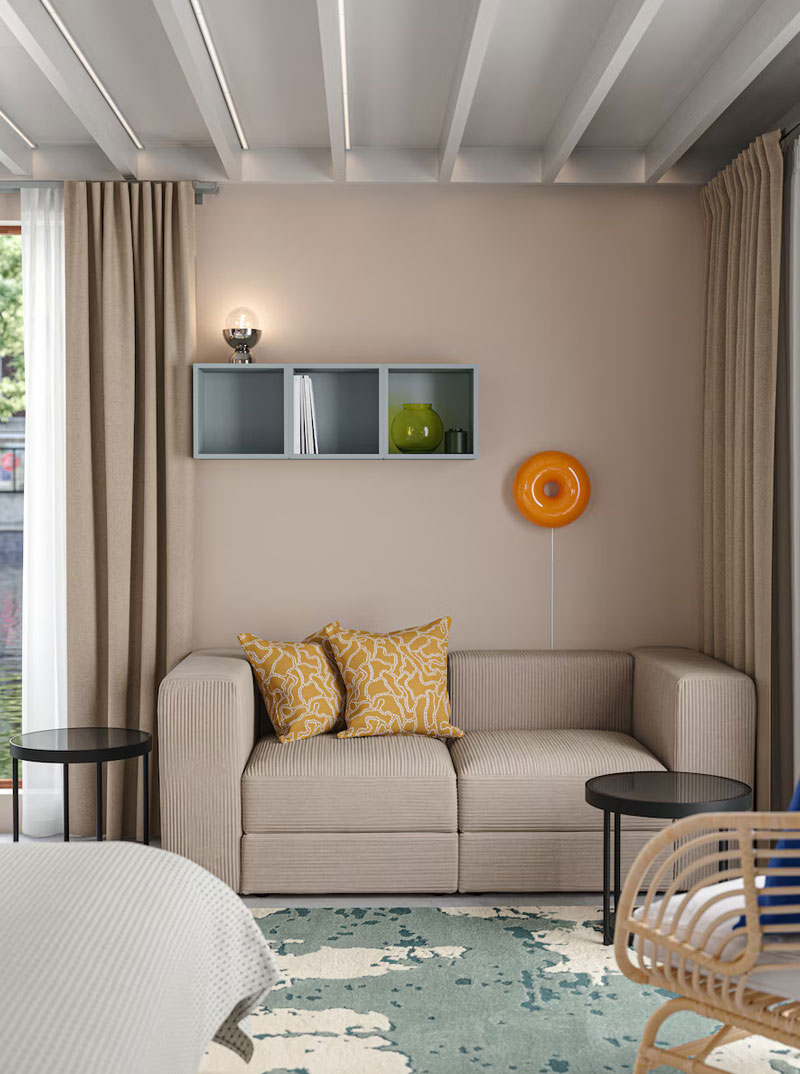 Un salón moderno, con un sofá modular beige con dos cojines amarillos estampados, una lámpara en forma de donut en la pared y un armario en forma de caja de color turquesa pálido.