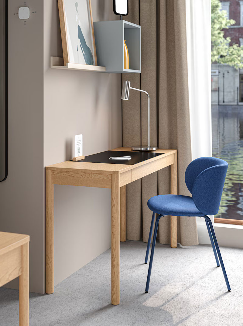 Un escritorio tipo madera, en una esquina de una habitación sobre una moderna lámpara de mesa, y en la pared una habitación junto a una estantería modular, hay una moderna silla azul con patas de metal.
