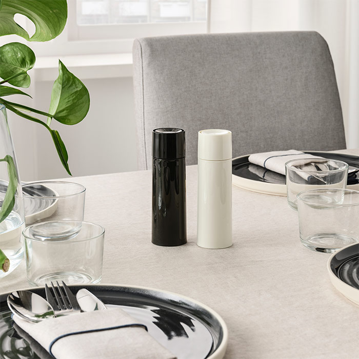 Mesa de comedor con molinillos blanco/negro, platos oscuros, vasos transparentes, cubiertos y una planta verde. Silla gris.